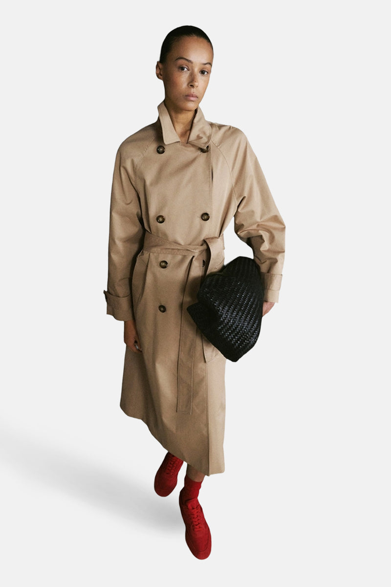 Veste longue beige de MANGO, avec ceinture et boutons marron, portée avec un sac noir tressé et des chaussures rouges.