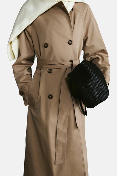 Veste longue beige de MANGO, avec ceinture nouée et poches, portée avec un pull blanc.
