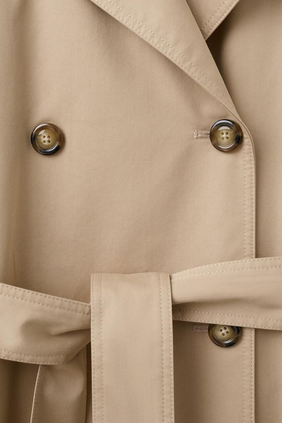 Veste longue beige de MANGO, avec une ceinture ton sur ton et des boutons marrons.
