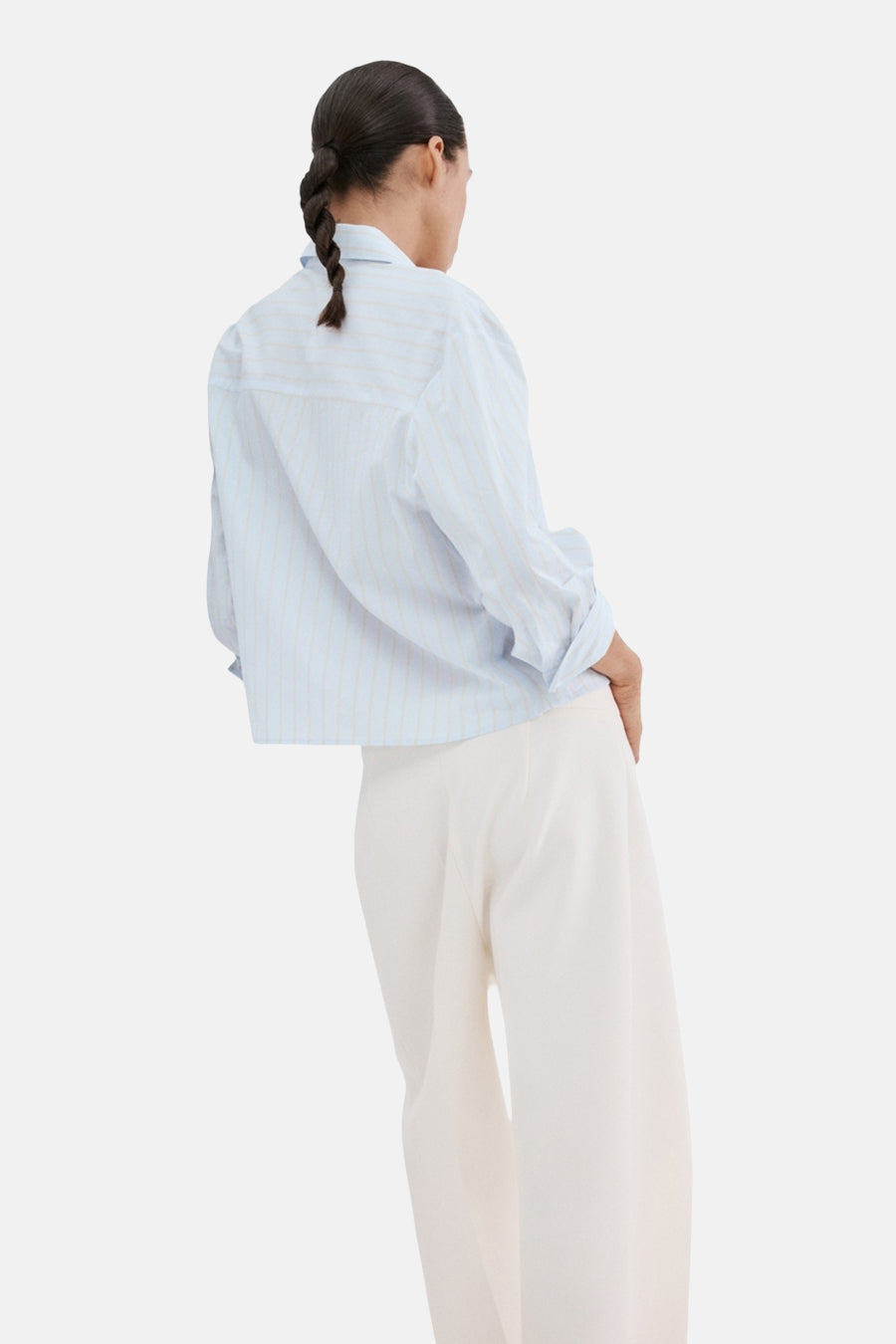 Blauw gestreept overhemd met lange mouwen van MANGO, gedragen met een witte broek en gevlochten kapsel, van achteren gezien.