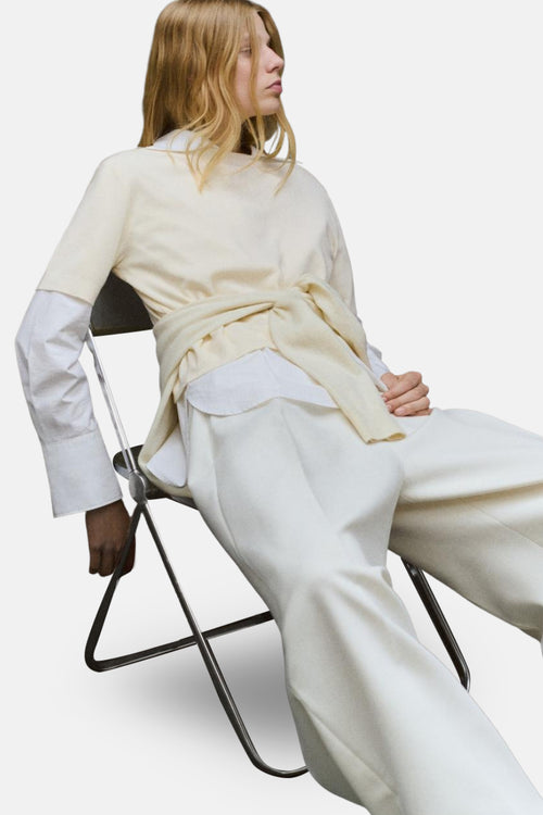 Pull à col rond écru de MANGO, porté avec une chemise blanche et un pantalon large assorti, femme assise sur une chaise.
