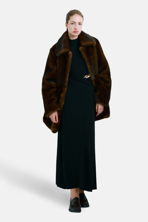 Robe noire de MANGO, longue et fluide avec une épingle dorée et portée avec un manteau de fourrure marron.
