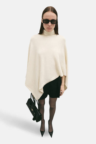 Ecru poncho van MANGO, gedragen over een zwarte jurk met panty en hoge hakken.