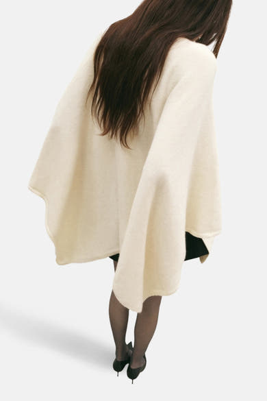 Poncho écru de MANGO, porté sur une robe noire avec des collants et des talons hauts.