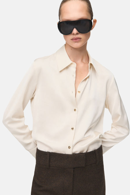 Chemise écrue à manches longues de MANGO, portée avec un pantalon marron et des lunettes de soleil noires.
