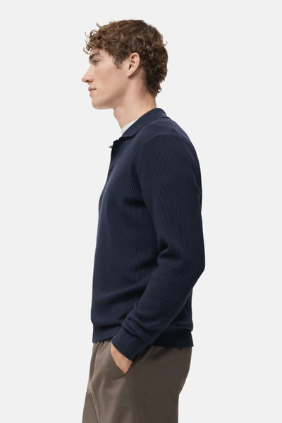Pull à col en V bleu de MANGO, en maille côtelée, porté avec un pantalon décontracté marron, vue de profil.