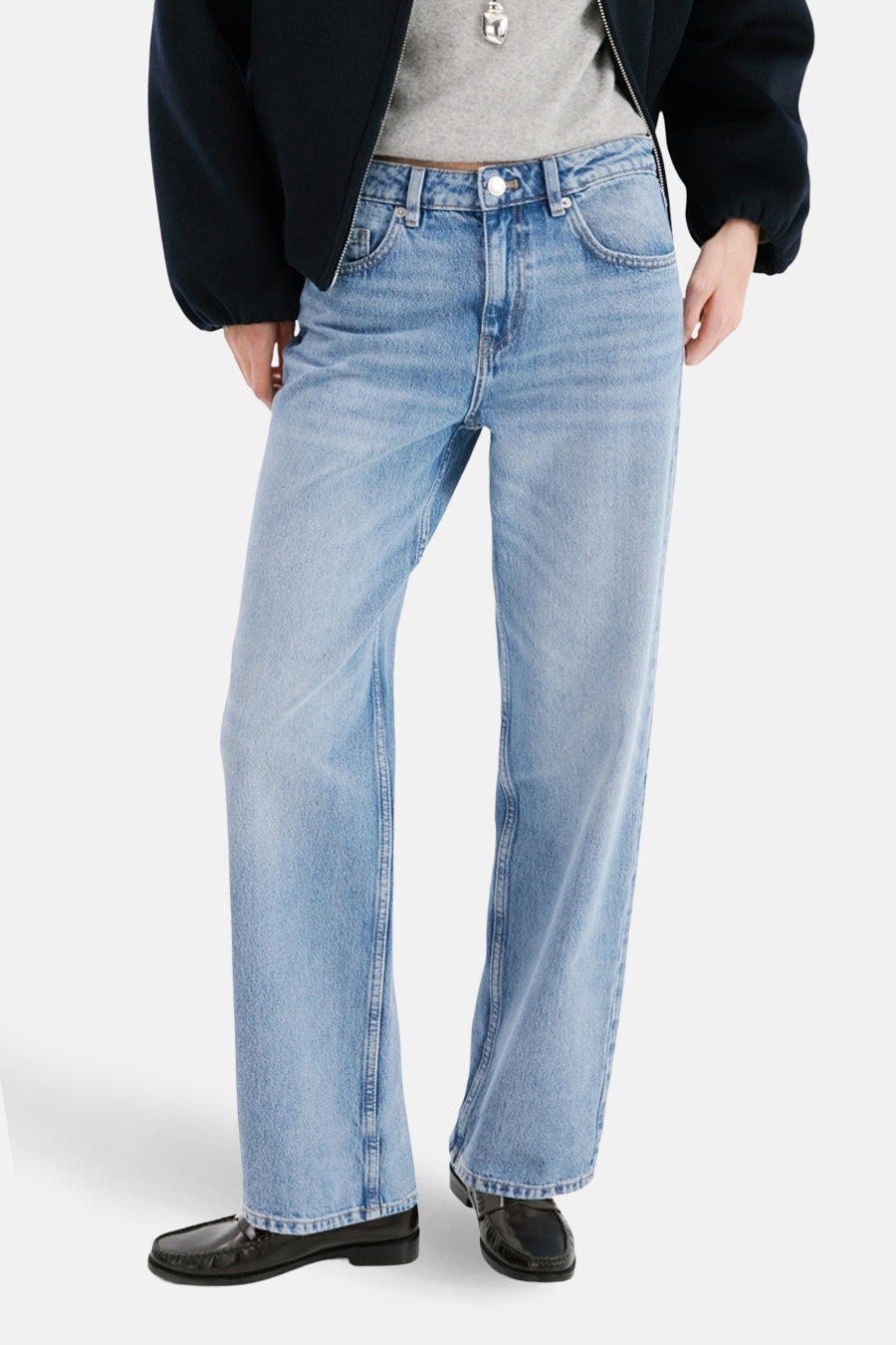 Jean droit en denim bleu clair de MANGO, porté avec un t-shirt gris, une veste bleu marine et des mocassins noirs.
