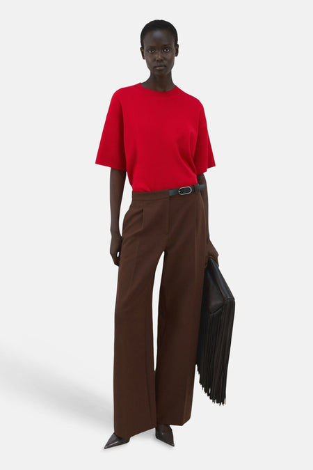 Pull col rond rouge de MANGO, porté avec un pantalon marron large, ceinture noire et un sac à franges.

