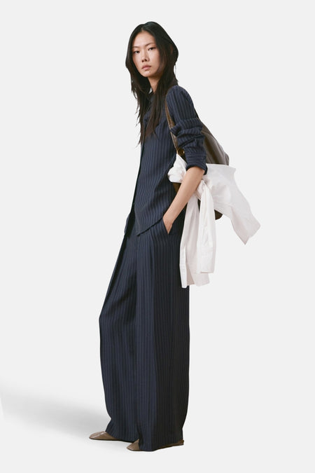 Pantalon bleu à rayures, de MANGO, porté avec une chemise blanche nouée et un sac à bandoulière marron, vue de côté.