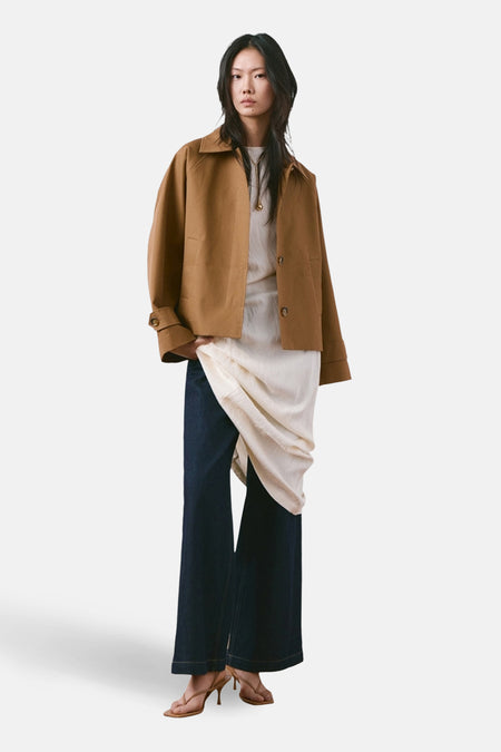 Veste courte marron de MANGO, portée avec un t-shirt long blanc cassé, un jean ample bleu foncé et des talons beige clair.