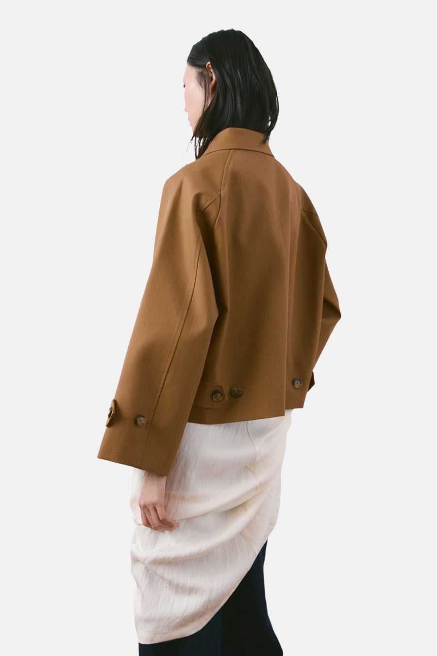 Veste courte brune de MANGO, avec des boutons, portée avec une jupe blanche drapée et un pantalon noir.