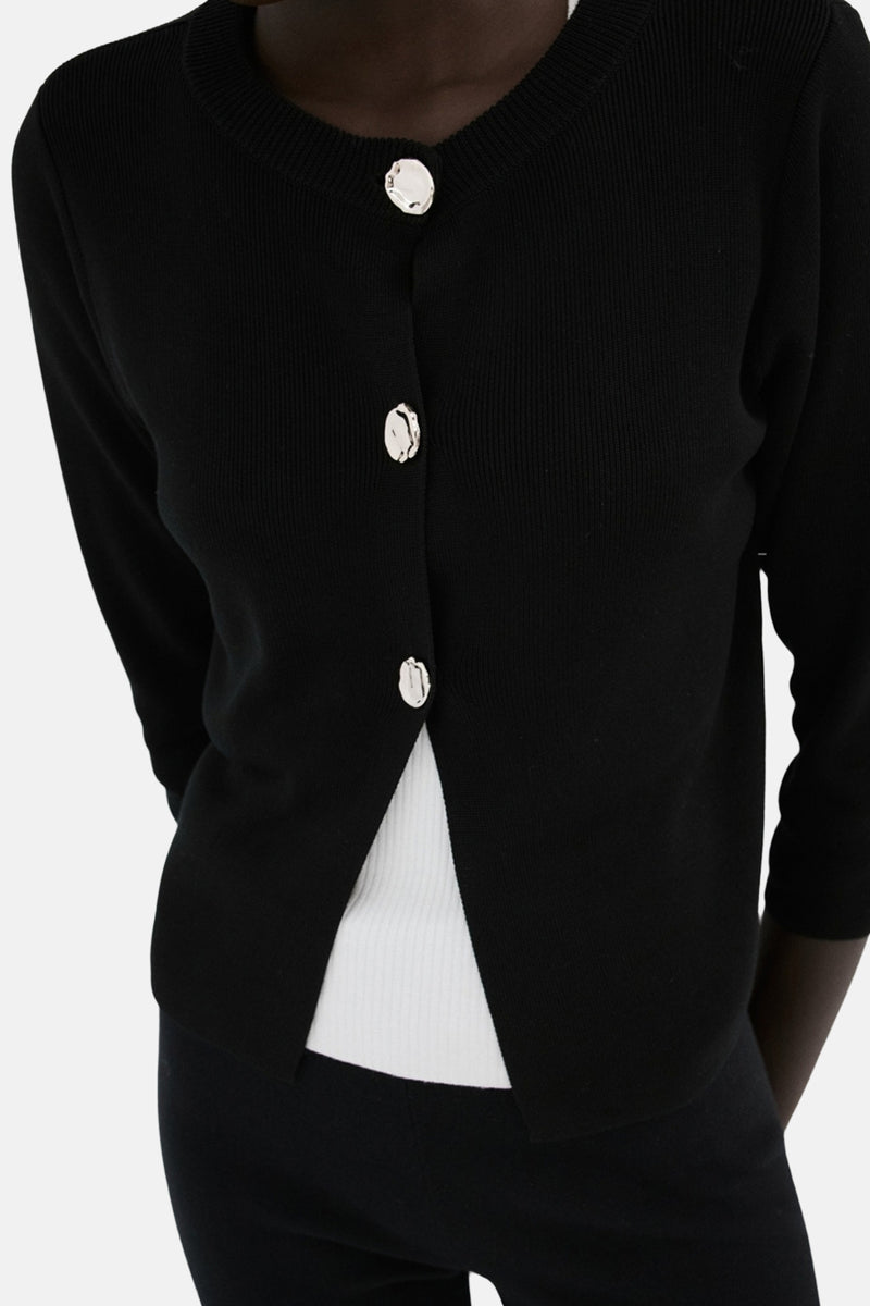 Zwarte MANGO-gilet, in fijne breiwerk met driekwartmouwen, gedragen over een witte top en een zwarte broek.