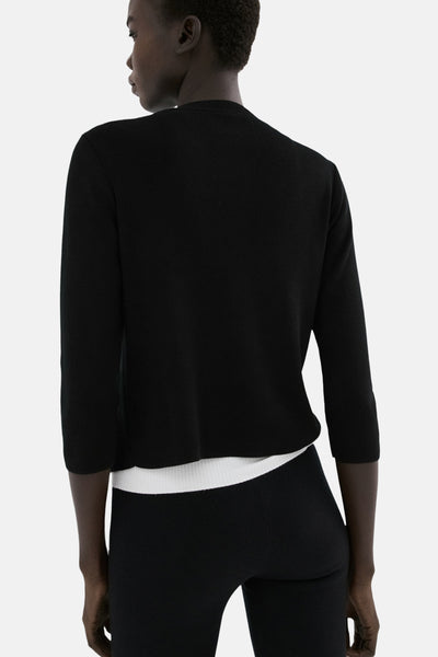 Gilet noir de MANGO, en maille fine avec manches trois-quarts, porté sur un haut blanc et un pantalon noir.