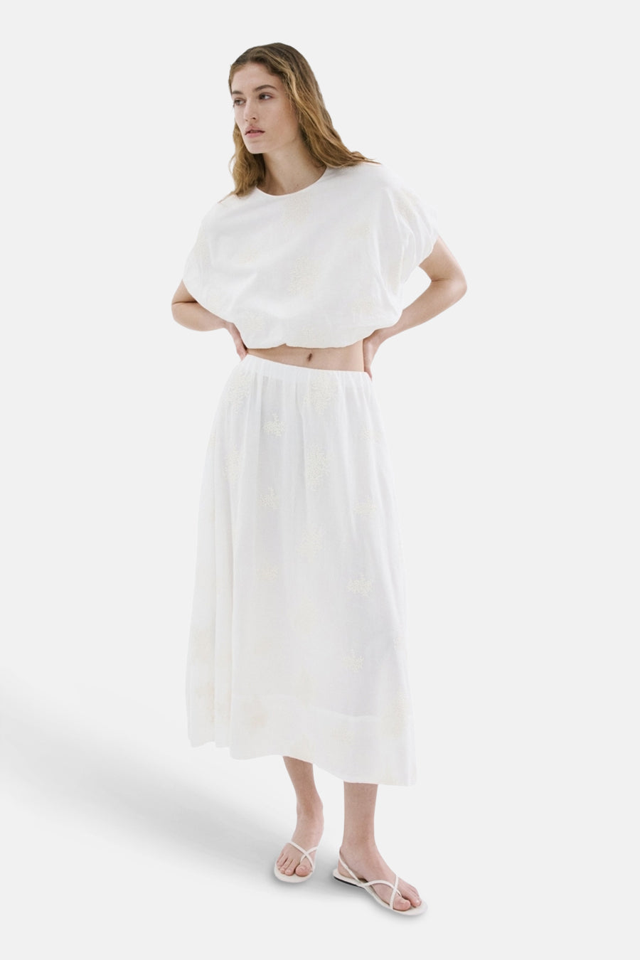 Jupe longue beige de MANGO, avec broderies florales délicates et fendue, portée avec un crop top assorti et des sandales blanches à brides.