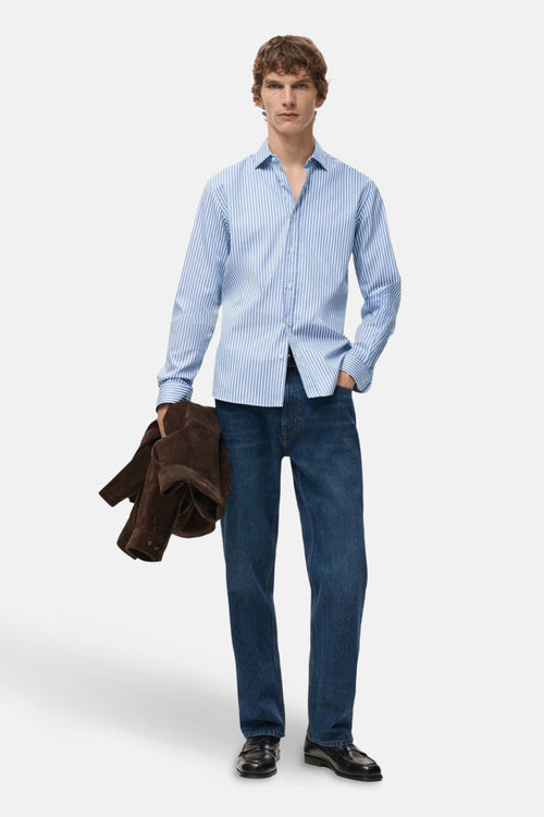 Chemise à manches longues bleue rayée de MANGO, portée avec un jean et des mocassins noirs, tenant une veste marron.
