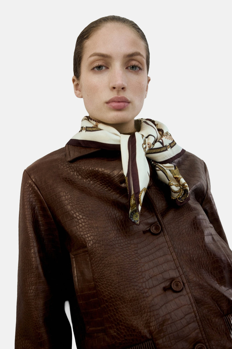 Veste courte brune en cuir imitation croco de MANGO, avec un foulard à motifs autour du cou.
