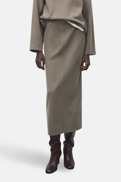 Lange bruine rok van MANGO, met een split aan de achterkant, gedragen met een witte crop top en bruine leren laarzen.
