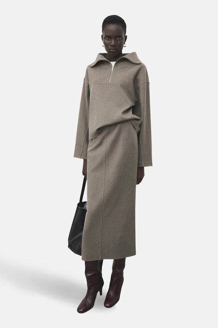 Jupe longue marron de MANGO, portée avec un pull zippé ton sur ton, un sac noir et des bottes marron.