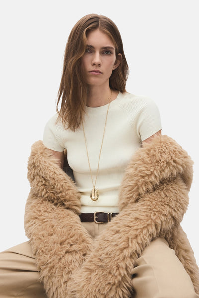 Collier long doré de MANGO, porté avec un pull côtelé et un manteau en fausse fourrure.
