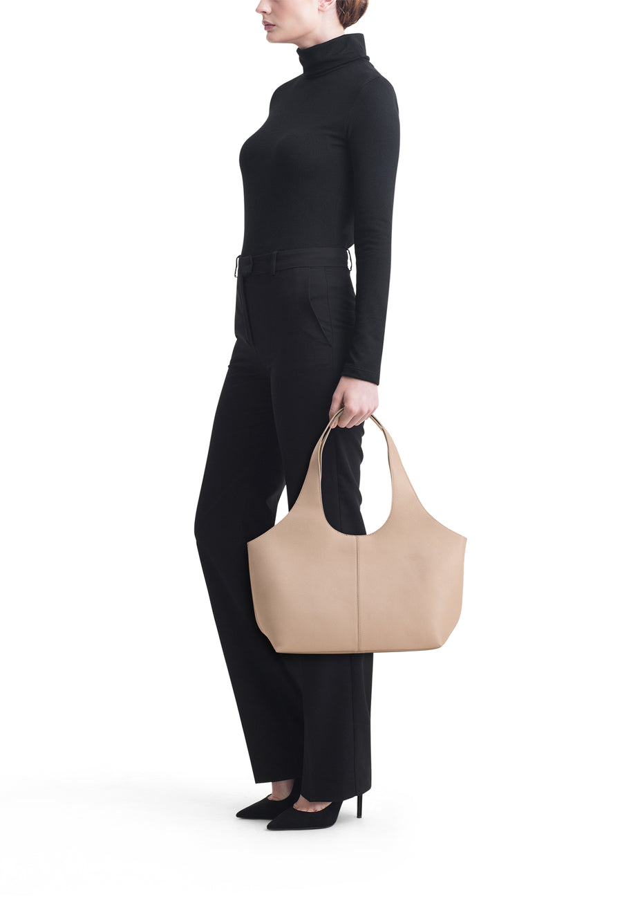 Beige schoudertas van MANGO, in de hand gedragen door een model in een zwarte broek en trui, op een witte achtergrond.