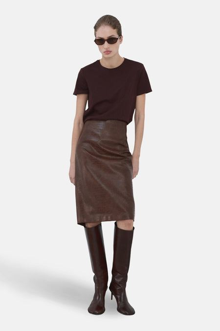 T-shirt à manches courtes bordeaux de MANGO, porté avec une jupe marron effet peau de serpent et des bottes hautes.