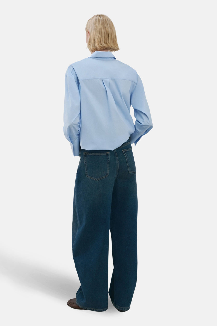 Chemise à manches longues bleue de MANGO, avec des plis au dos, portée avec un jean ample et des chaussures marron.