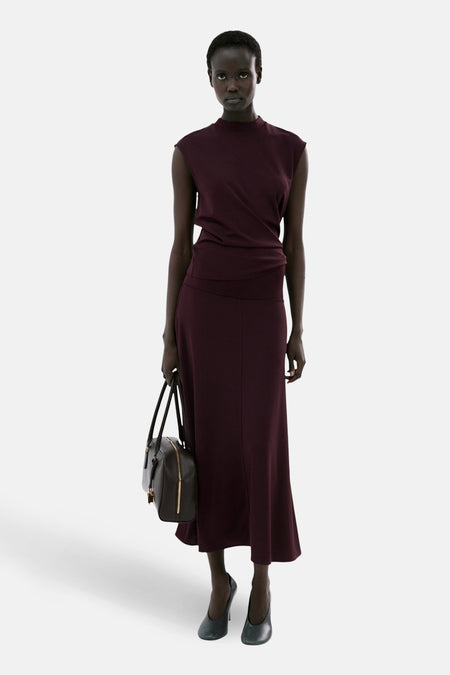 Top sans manches bordeaux par MANGO, porté avec une jupe longue et des escarpins noirs, tenant un sac à main marron.
