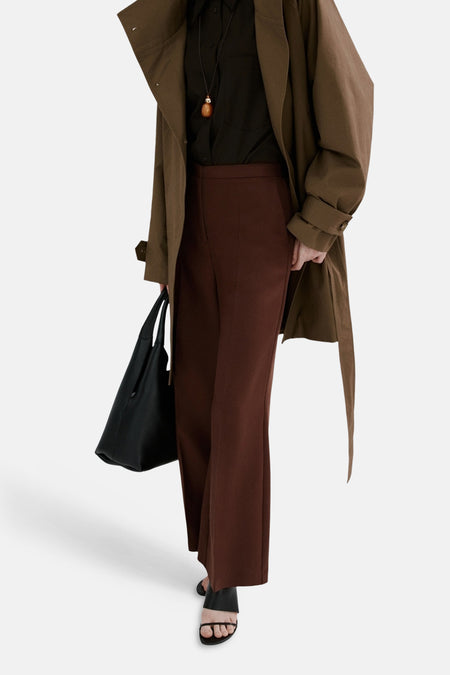 Pantalon brun de MANGO, coupe droite, porté avec un chemisier foncé, un trench marron et des sandales noires.