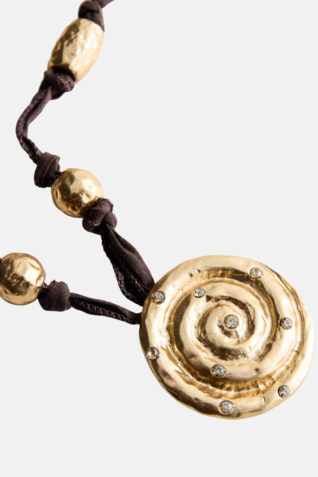 Close-up van een bruine ketting van MANGO met een ronde, gehamerde gouden hanger, spiraalmotief en strass.
