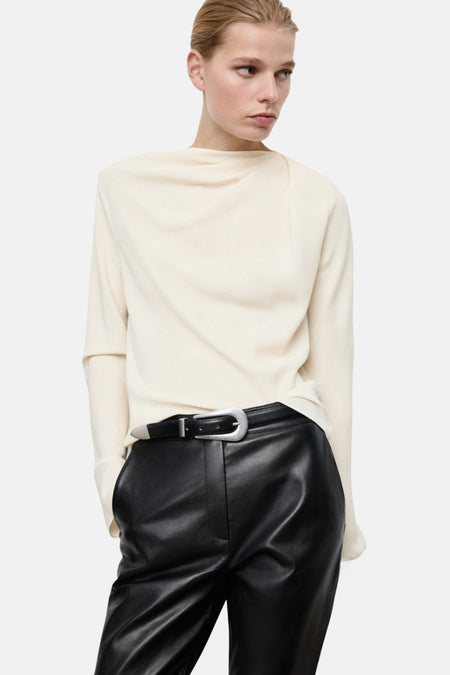 Blouse à manches longues écru de MANGO, avec un drapé asymétrique sur le devant et portée avec un pantalon en cuir noir.
