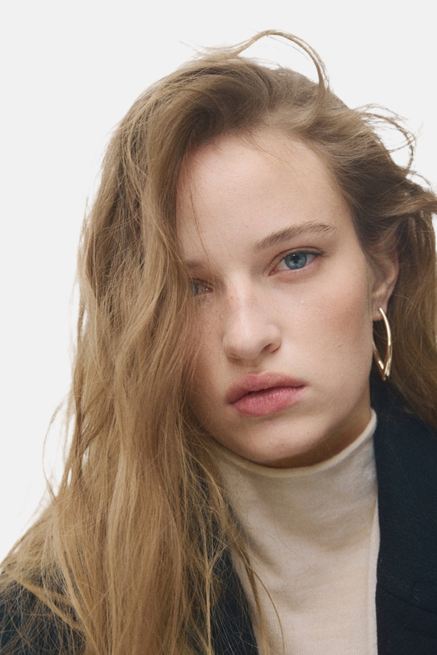 Boucles d'oreilles dorées de MANGO, portées par une femme avec un pull col roulé beige et une veste noire.