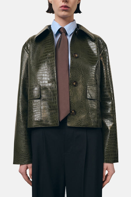 Veste courte kaki par MANGO, en similicuir imprimé façon crocodile, portée sur une chemise rayée et une cravate marron.
