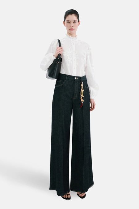 Chemisier blanc à manches longues de MANGO, orné de broderies florales, porté avec un jean taille haute.
