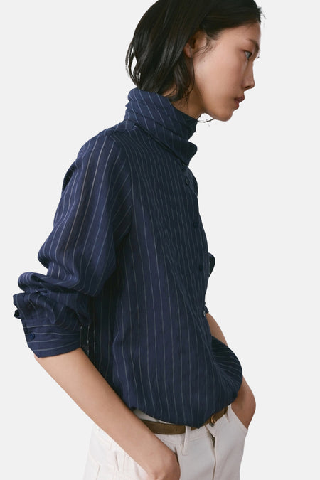Chemisier bleu à manches longues de MANGO, rayures verticales blanches, col montant, porté avec un pantalon blanc et une ceinture marron, vue de côté.