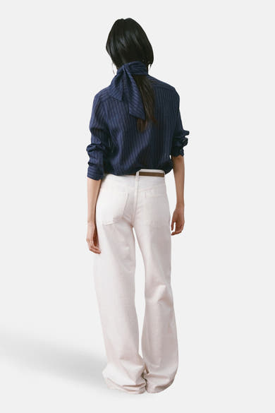 Blouse à manches longues bleue à rayures by MANGO, avec un nœud au dos, portée avec un pantalon blanc et une ceinture marron.