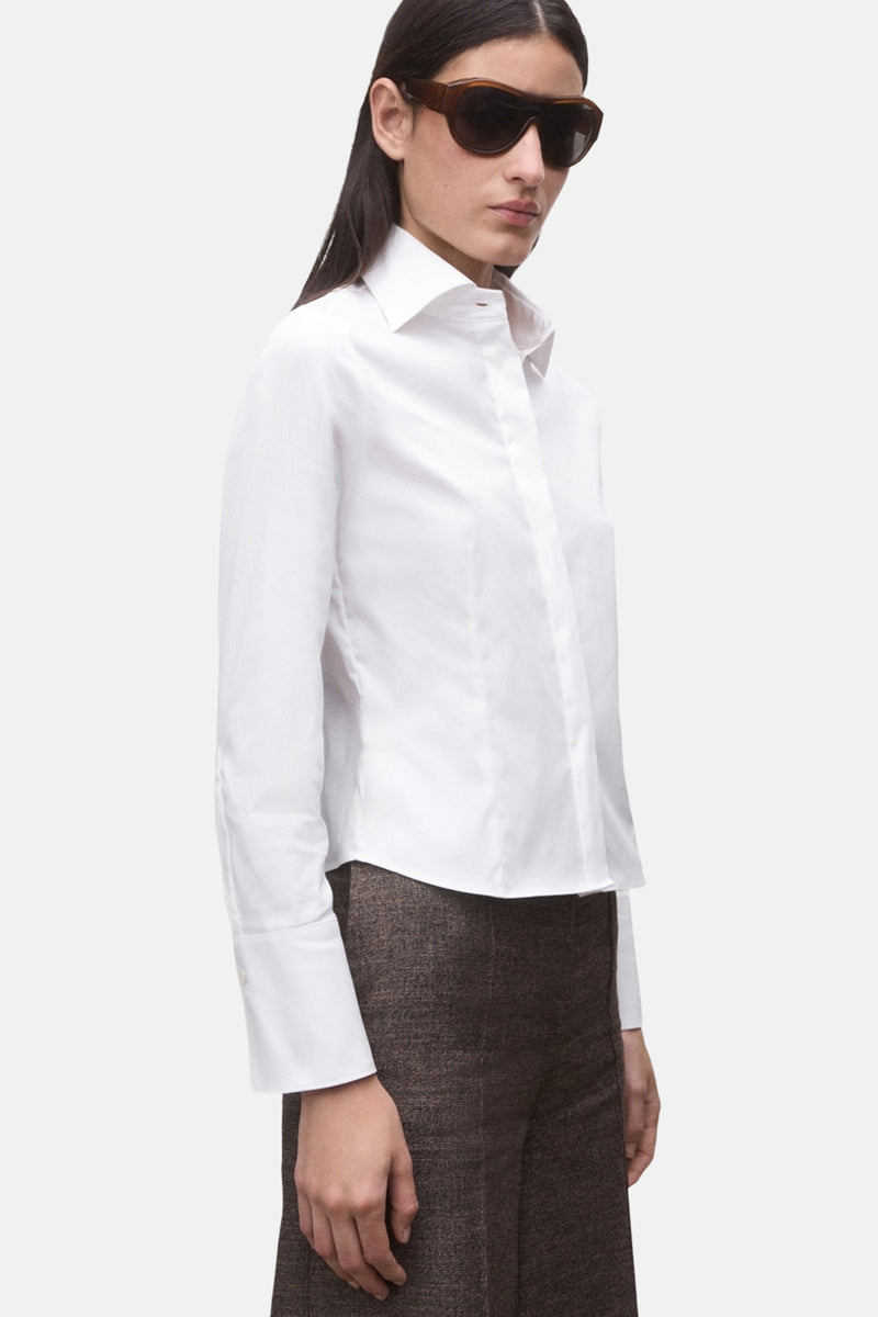 Chemise à manches longues blanche de MANGO, portée avec un pantalon marron texturé et des lunettes de soleil.