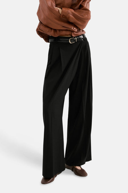 Pantalon noir de MANGO, ample et fluide, porté avec une ceinture noire et une veste en cuir marron.
