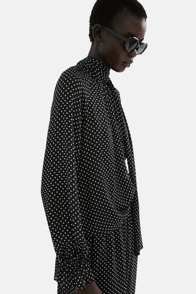 Chemisier noir à pois blancs de MANGO, avec col montant à nouer et manches longues froncées.