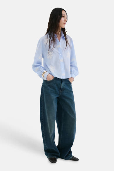 Blauwe blouse met lange mouwen van MANGO, versierd met witte bloemenborduursels, gedragen met een wijde spijkerbroek.