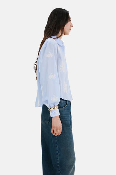 Blauwe blouse met lange mouwen van MANGO, versierd met witte bloemenborduursels, gedragen met een gouden armband en een blauwe spijkerbroek.