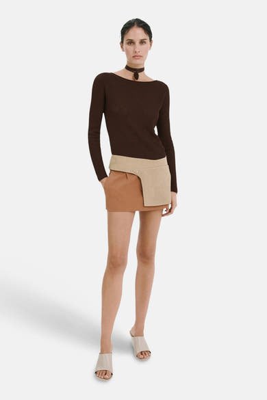 Pull brun en maille côtelée de MANGO à col bateau, porté avec une mini-jupe bicolore et des mules claires.