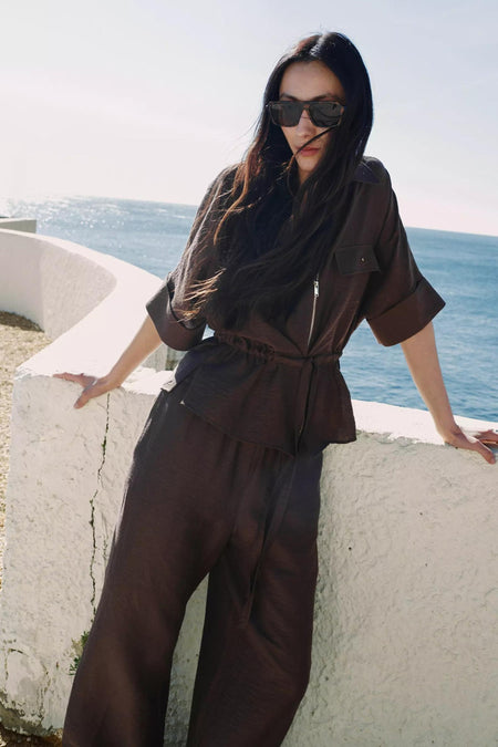 Blouse à manches courtes brune de MANGO, avec fermeture éclair, poches poitrine et taille coulissée, portée avec un pantalon assorti.