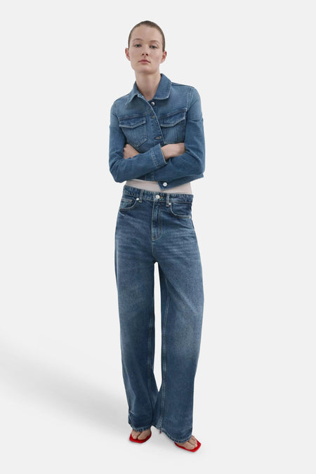 Veste en jean denim bleu de MANGO, courte, portée par un mannequin aux bras croisés avec un jean large et des sandales rouges.
