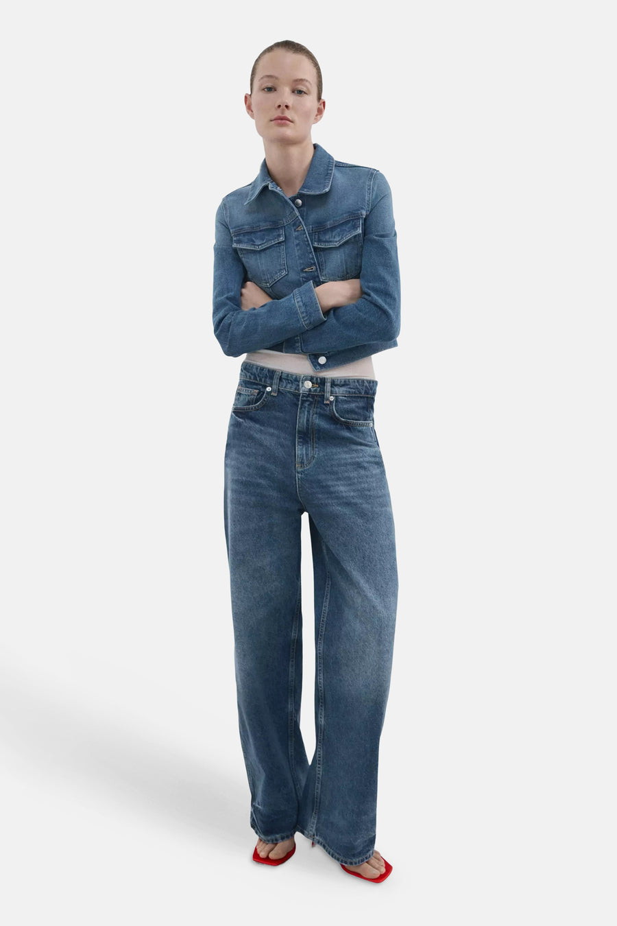 Korte blauwe denim spijkerjas van MANGO, gedragen door een model met gekruiste armen, een wijde spijkerbroek en rode sandalen.