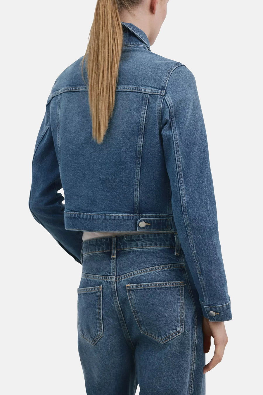 Veste en jean bleue de MANGO, coupe courte, portée de dos avec un jean assorti et des boutons métalliques visibles.