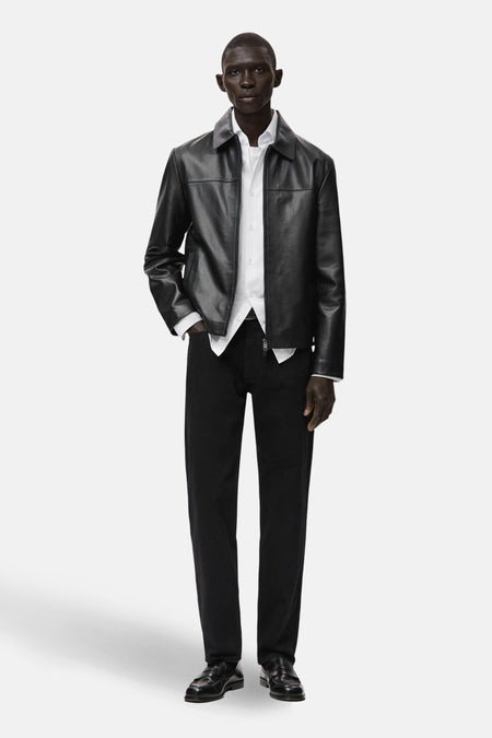 Veste courte noire en cuir, de MANGO, portée sur une chemise blanche et un pantalon noir, avec des chaussures en cuir assorties.
