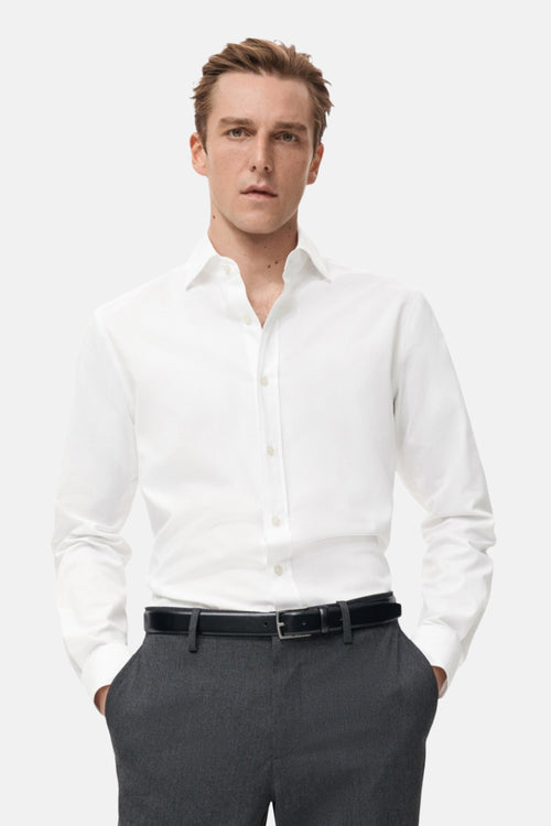 Chemise blanche à manches longues de MANGO, portée avec un pantalon gris et une ceinture noire.
