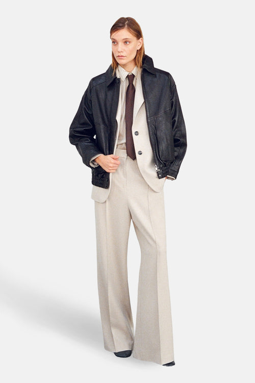 Pantalon beige de MANGO, coupe évasée, porté avec une veste en cuir noire et une chemise rayée.
