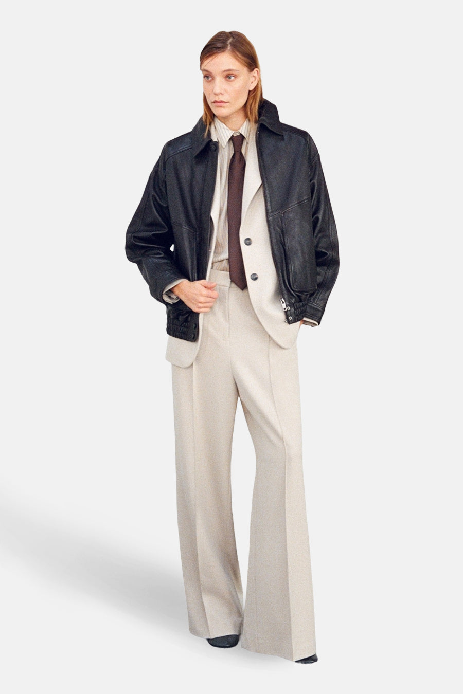 Pantalon beige de MANGO, coupe évasée, porté avec une veste en cuir noire et une chemise rayée.
