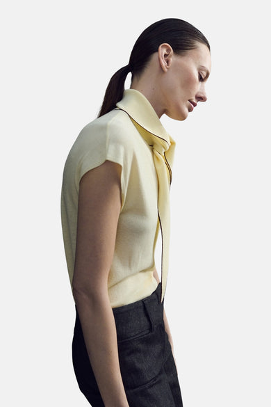Chemisier jaune sans manches de MANGO, avec col montant et finitions contrastantes marron, porté avec un pantalon foncé.
